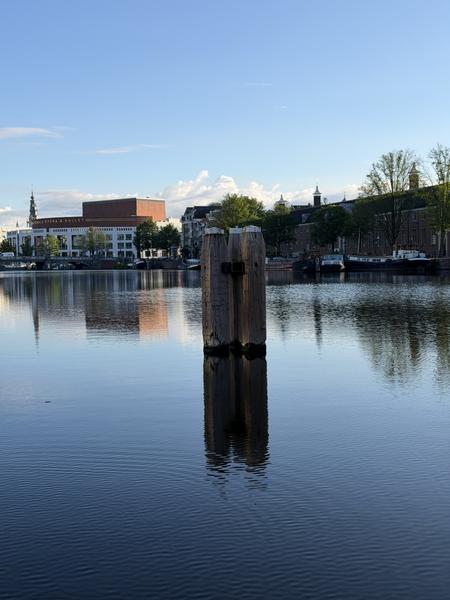 This tranquil photo captures a serene morning view of the Amstel in Centrum, Amsterdam, with a prominent wooden post reflecting in the calm water